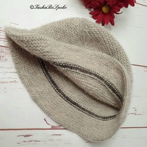 May include: A beige knitted hat with two dark brown stripes. The hat is made of a soft, textured yarn and is folded over. The background includes red flowers and the text "TashaBeSpoke".