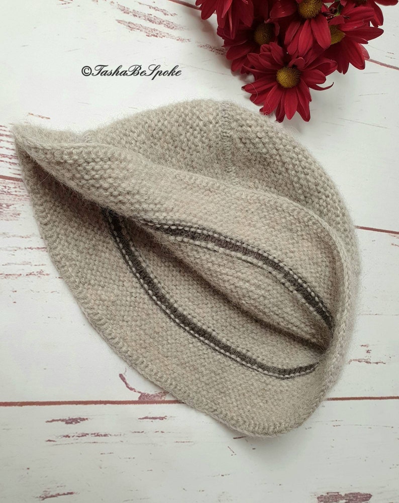 May include: A beige knitted hat with two dark brown stripes. The hat is made of a soft, textured yarn and is folded over. The background includes red flowers and the text "TashaBeSpoke".