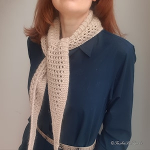 Puede incluir: Una bufanda de ganchillo beige con un delicado patr&oacute;n de calado. La bufanda est&aacute; drapeada alrededor del cuello de una persona que lleva un vestido azul oscuro.