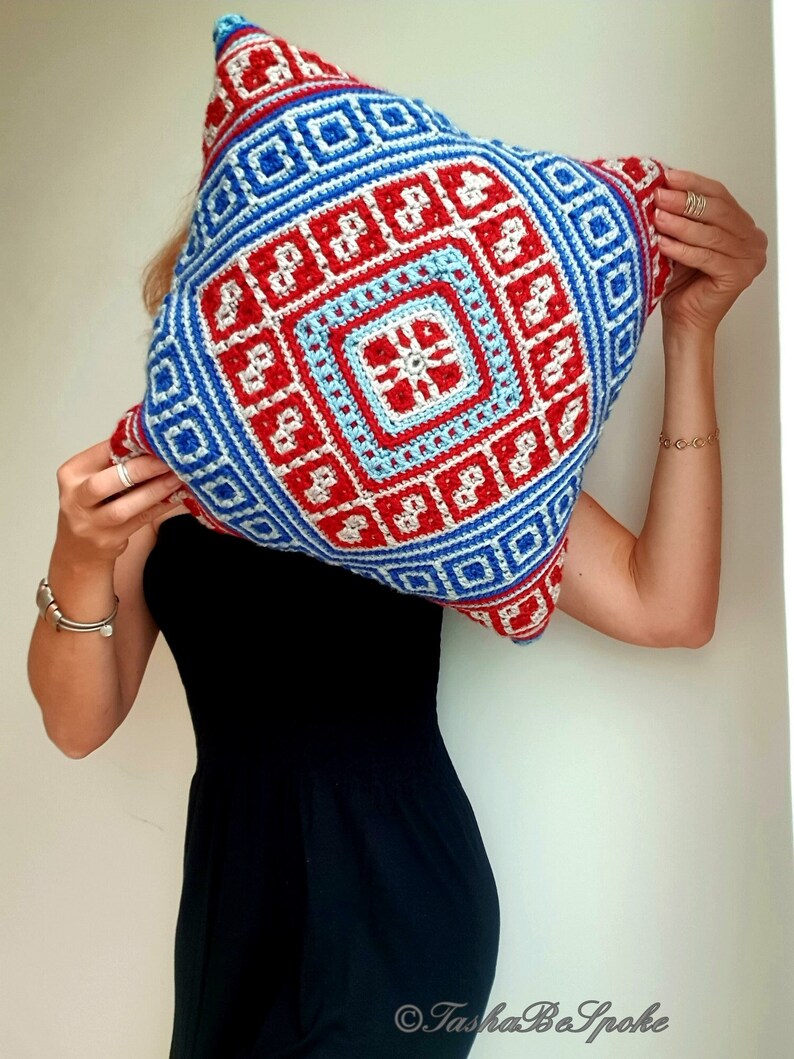 Op de afbeelding: Een vierkant gehaakt kussen met een geometrisch ontwerp in rood, wit en blauw. Het ontwerp heeft een centraal stervormig patroon, omgeven door concentrische vierkanten en rechthoeken. Het kussen wordt door een persoon vastgehouden.