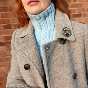 May include: A woman wearing a light blue turtleneck sweater under a grey wool coat. The coat has a large, colourful brooch pinned to the lapel.