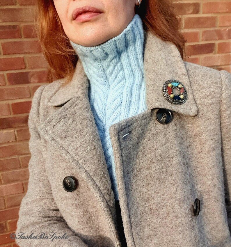 May include: A woman wearing a light blue turtleneck sweater under a grey wool coat. The coat has a large, colourful brooch pinned to the lapel.