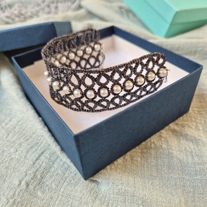 Puede incluir: Un brazalete de encaje negro con detalles de perlas blancas, presentado en una caja azul. El brazalete es delicado e intrincado, con un dise&ntilde;o tejido.