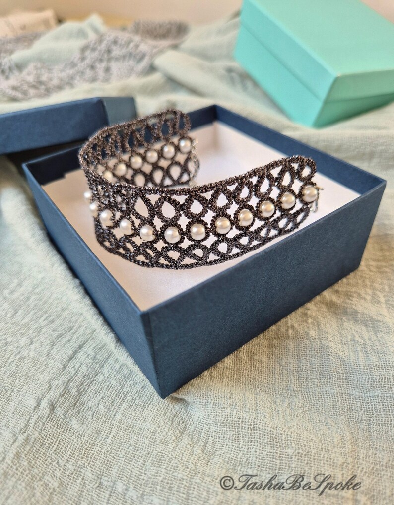 Puede incluir: Un brazalete de encaje negro con detalles de perlas blancas, presentado en una caja azul. El brazalete es delicado e intrincado, con un dise&ntilde;o tejido.