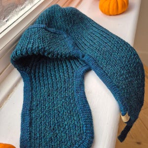 K&ouml;nnte beinhalten: Eine tealblaue Strickm&uuml;tze mit einer langen, spitzen Kapuze. Die M&uuml;tze ist aus dickem, strukturiertem Garn gefertigt und hat ein schlichtes, klassisches Design.