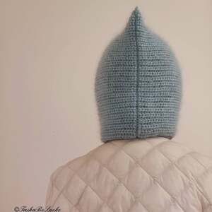 Pu&ograve; includere: Un cappello all'uncinetto blu chiaro a punta. Il cappello &egrave; realizzato con un filato morbido e strutturato e viene indossato sopra una giacca trapuntata bianca. Il design del cappello &egrave; semplice, con una parte superiore a punta e una forma aderente.