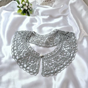 May include: A gray crocheted collar with a pearl button in the center. The collar is laying on a white satin fabric.