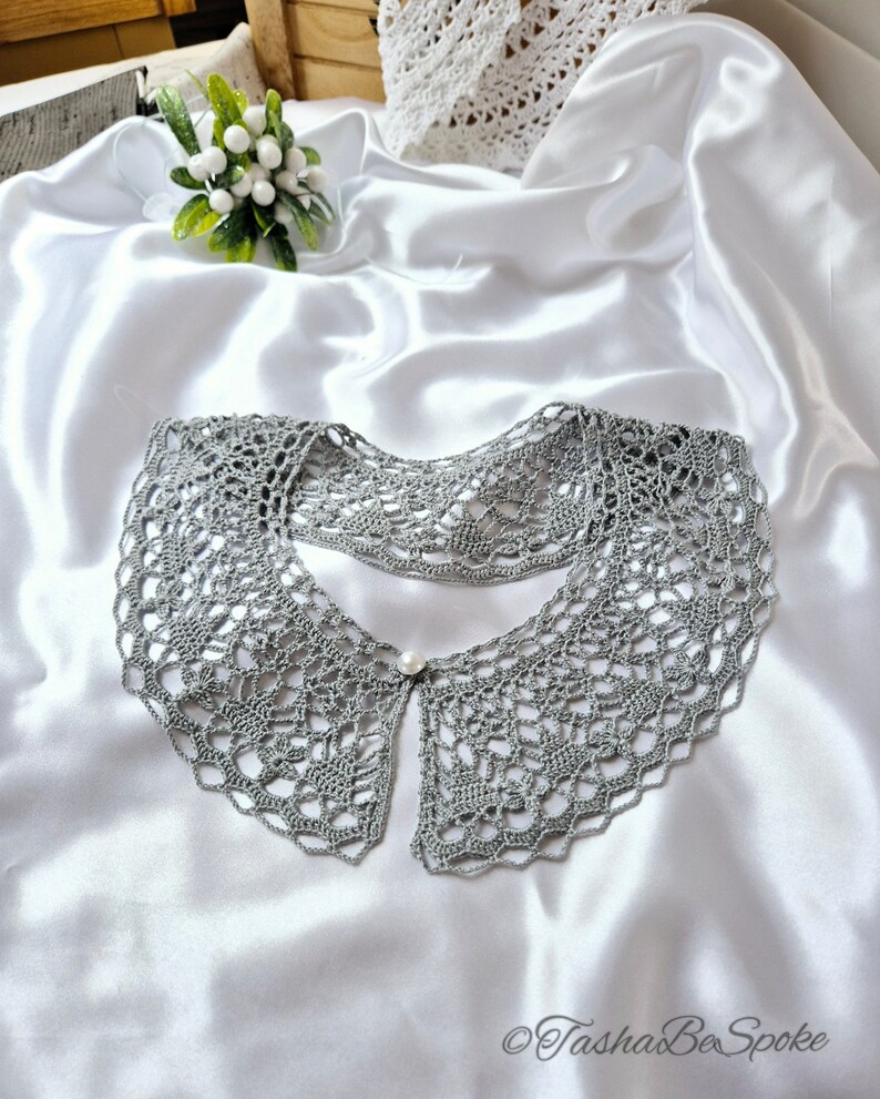 May include: A gray crocheted collar with a pearl button in the center. The collar is laying on a white satin fabric.