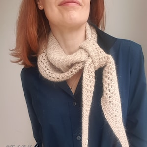 Puede incluir: Una bufanda de punto beige con un delicado patr&oacute;n de encaje. La bufanda est&aacute; atada alrededor del cuello de una persona que lleva una camisa azul oscuro.