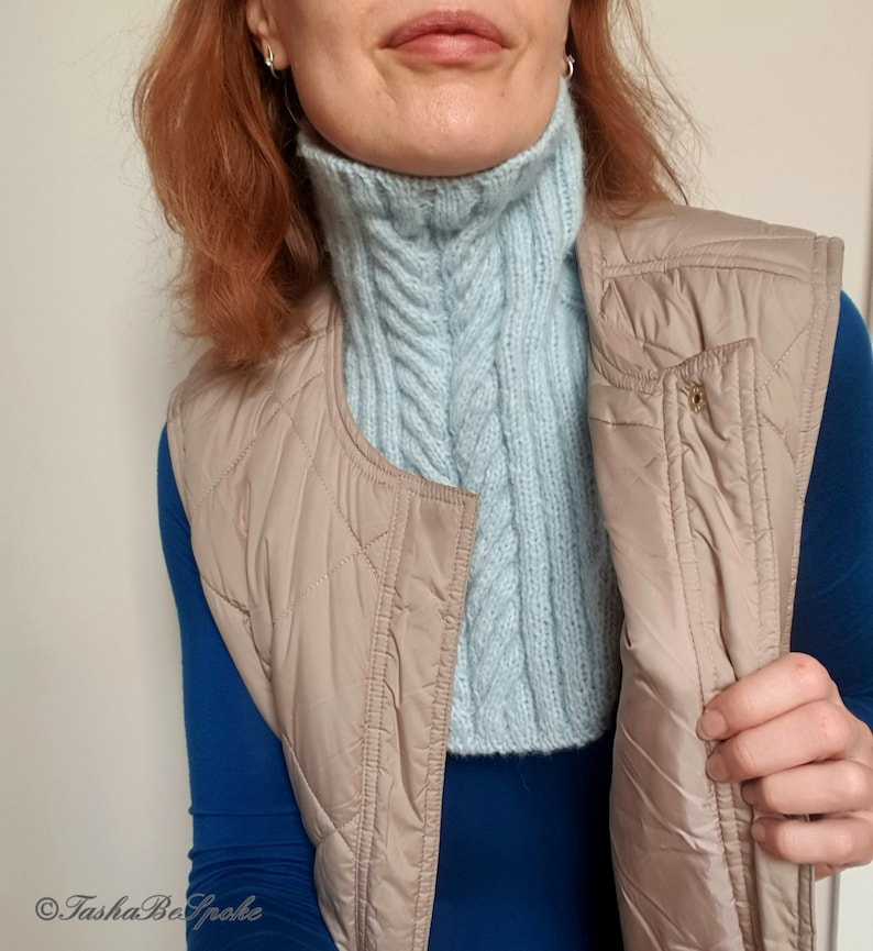 May include: A light blue knitted turtleneck collar with a cable knit pattern. The collar is worn over a blue long-sleeved shirt and a beige quilted vest.