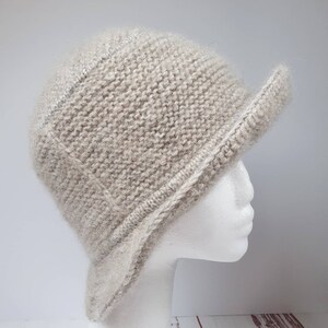 May include: A light beige knitted hat with a short brim, displayed on a white mannequin head. The hat has a textured appearance and a structured design, suitable for cooler weather.