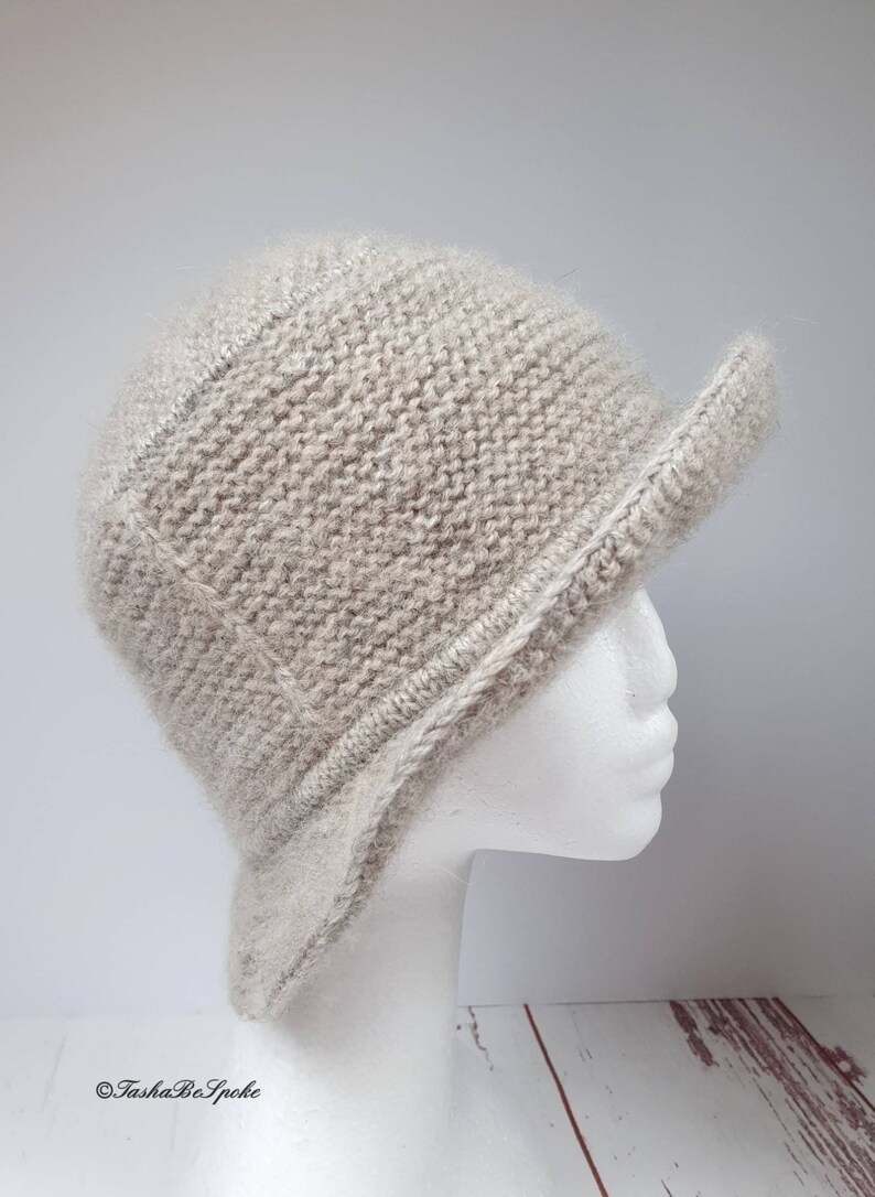 May include: A light beige knitted hat with a short brim, displayed on a white mannequin head. The hat has a textured appearance and a structured design, suitable for cooler weather.