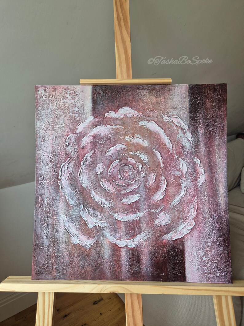 May include: A square canvas artwork depicting a textured rose in pink and white, displayed on a wooden easel. The rose has a raised relief effect. The artist's signature, "TashaBeSpoke", is visible in the upper right corner of the artwork.