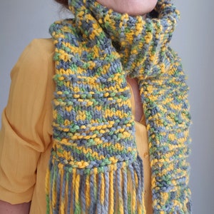 May include: A knitted scarf with a striped pattern in shades of gray, yellow, and green. The scarf has long fringe tassels.
