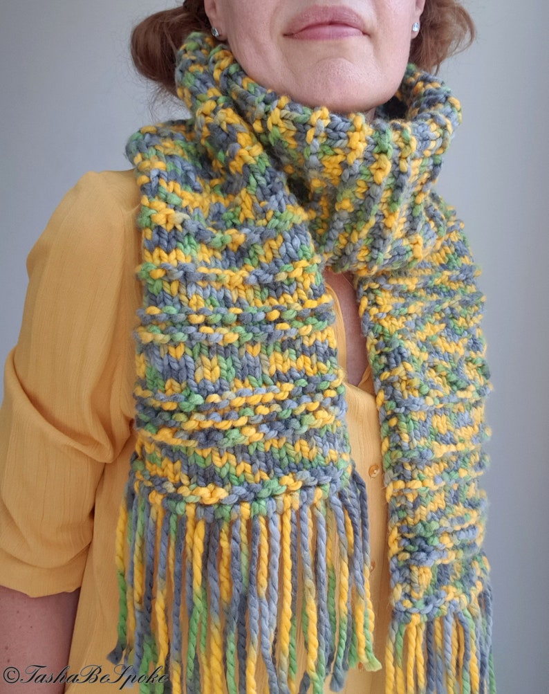 May include: A knitted scarf with a striped pattern in shades of gray, yellow, and green. The scarf has long fringe tassels.