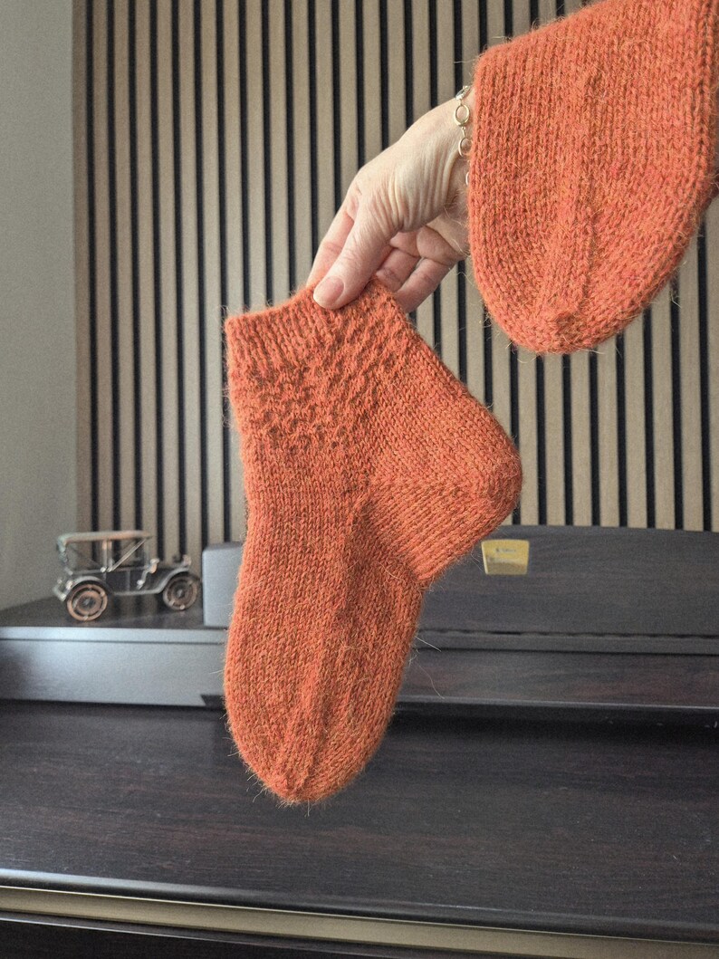 May include: A pair of orange knitted socks, one held up by a hand, with a matching knitted sleeve visible. The socks have a textured pattern on the top. The background includes a wooden slatted wall and a dark wooden surface.