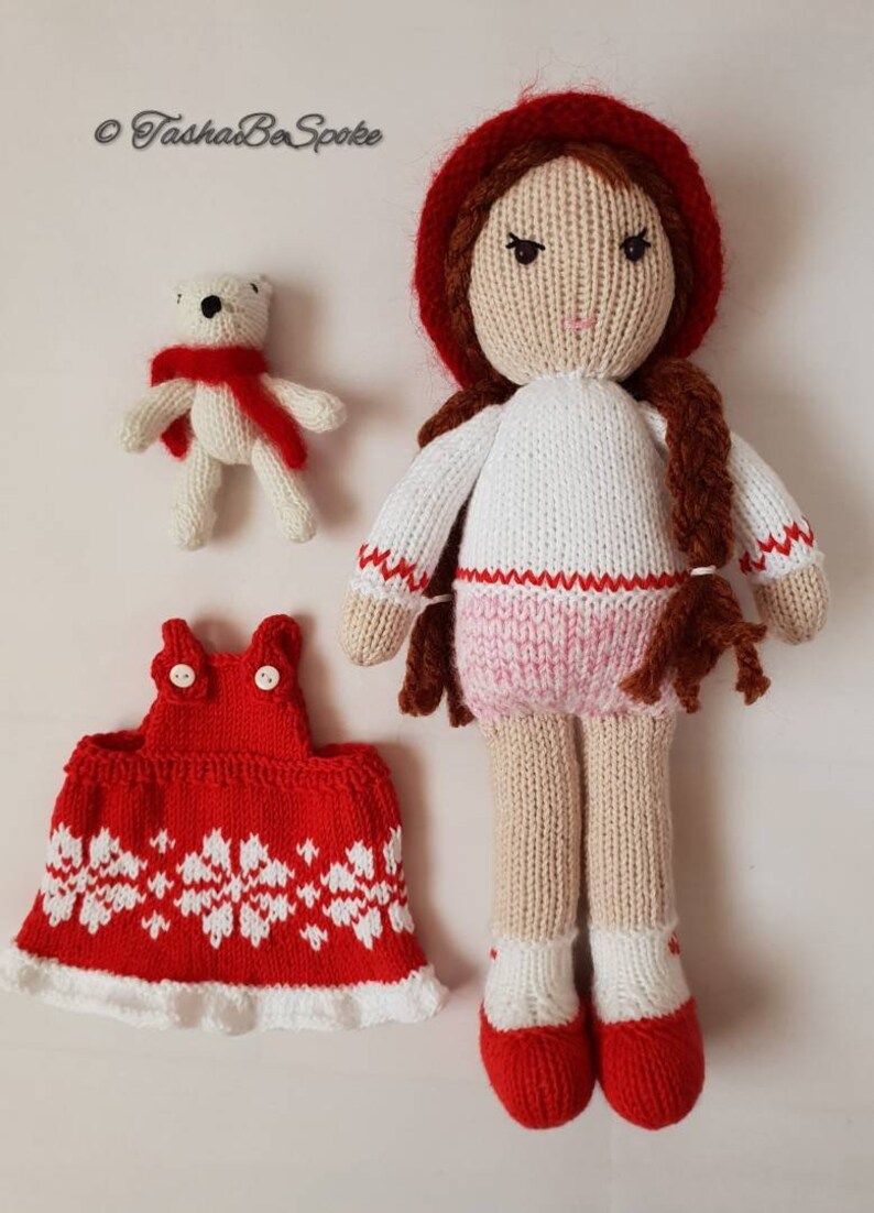 May include: Knitted doll with red hat, white sweater, pink skirt, and red shoes. The doll is holding a white knitted teddy bear wearing a red scarf. The doll also has a red knitted dress with white snowflake pattern.