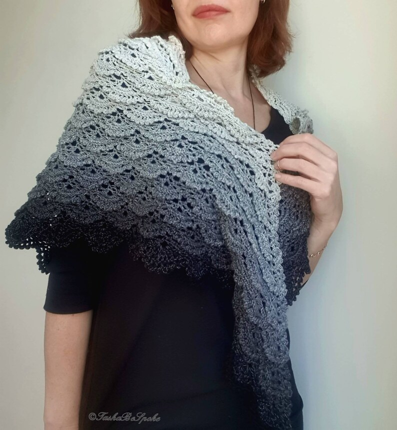 May include: A grey and black ombre crochet shawl with a scalloped edge. The shawl is draped over a person's shoulders.