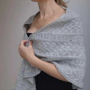 May include: A light gray knitted shawl with a delicate cable pattern. The shawl is draped over a person's shoulders and is worn with a black top.