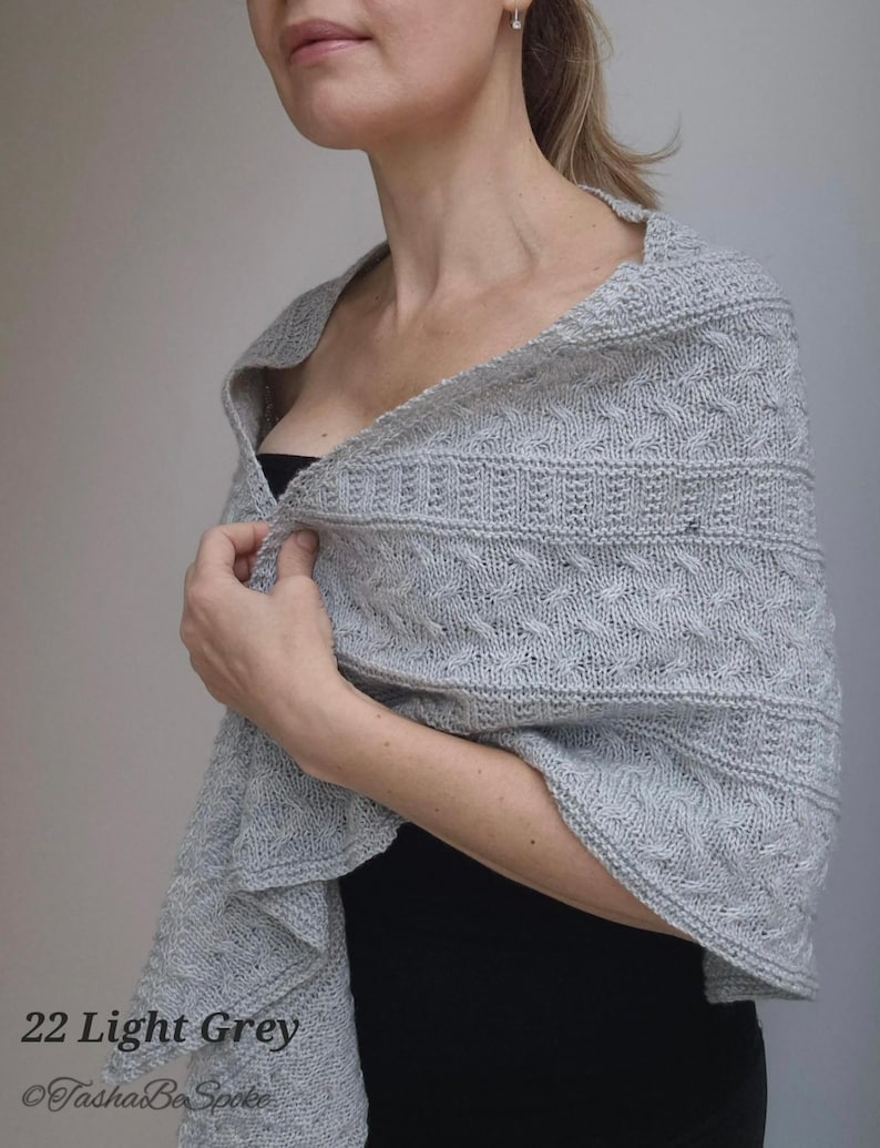 May include: A light gray knitted shawl with a delicate cable pattern. The shawl is draped over a person's shoulders and is worn with a black top.