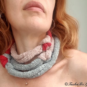 May include: A knitted cowl in shades of gray, pink, and red. The cowl is wrapped around the neck, showcasing a braided design. The handmade accessory is a cozy and stylish addition to any outfit.