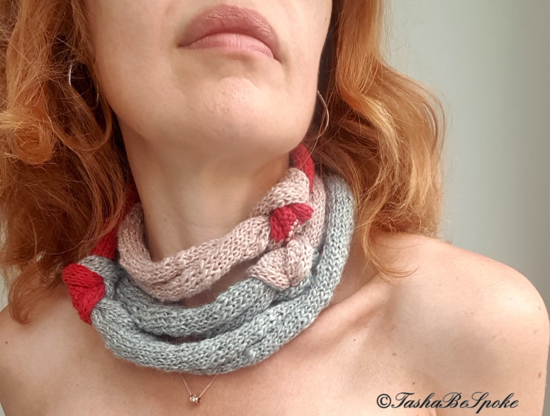 May include: A knitted cowl in shades of gray, pink, and red. The cowl is wrapped around the neck, showcasing a braided design. The handmade accessory is a cozy and stylish addition to any outfit.