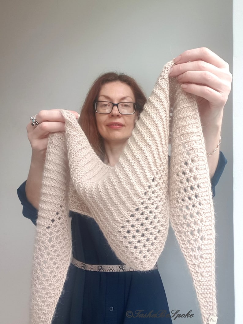 Puede incluir: Un chal de punto beige con un delicado patr&oacute;n de encaje. El chal lo sostiene una persona que lleva un vestido azul oscuro.
