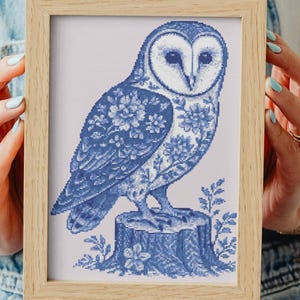 May include: Framed cross-stitch art featuring a blue and white owl design. The owl is perched on a tree stump, with floral patterns on its wings and body. The artwork is displayed in a light brown wooden frame.