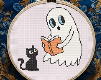 May include: A cross-stitch embroidery hoop featuring a white ghost reading a book with an orange cover, and a black cat. The background is light pink, and the hoop is held in a wooden frame. The background is a dark blue fabric with a gold floral pattern.
