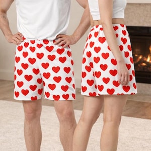 May include: A pair of white shorts featuring a repeating pattern of solid red hearts. The shorts have a relaxed fit and are worn with a plain white tank top. The fabric is white with evenly spaced red heart shapes across the entire garment.