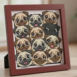May include: A framed cross-stitch artwork featuring a grid of pug faces in various shades of tan, brown, and black. The frame is a dark reddish-brown color. A small succulent in a white pot and a notebook with a pen are on the wooden surface.