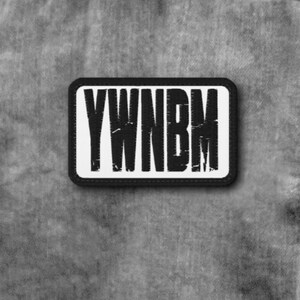 May include: A rectangular patch with a white background and a thick black border. The patch features the black text YWNB M in a distressed, vertical, sans-serif font. The patch is attached to a grey denim fabric with visible stitching and texture.