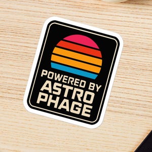 May include: A rectangular sticker with rounded corners featuring a black background, a stylized sunset graphic with horizontal stripes in red, orange, and blue, and the white text "POWERED BY ASTROPHAGE" printed in a bold, sans-serif font.