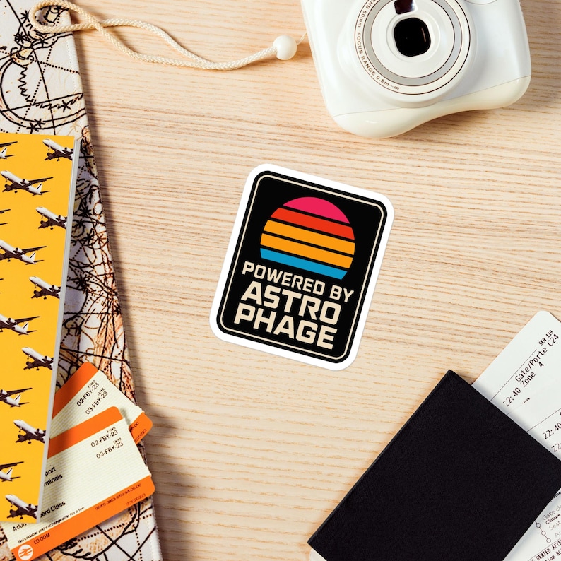 May include: A rectangular sticker with rounded corners featuring a black background, a stylized sunset graphic with horizontal stripes in red, orange, and blue, and the white text "POWERED BY ASTROPHAGE" printed in a bold, sans-serif font.