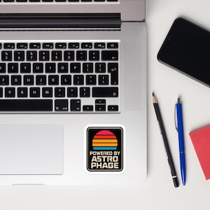 May include: A square sticker featuring a retro-style sunset graphic with horizontal stripes in red, orange, and yellow, positioned above the text "POWERED BY ASTROPHAGE" in a bold, sans-serif font, applied to the surface of a silver laptop.