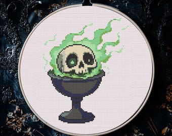 May include: Cross-stitch embroidery featuring a skull with green eyes and green flames rising from a dark cauldron. The design is set in a white fabric within a round wooden embroidery hoop, perfect for Halloween decor.