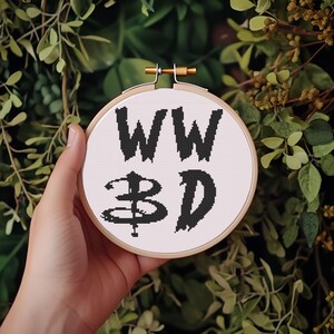 Könnte beinhalten: Ein Kreuzstich-Stickrahmen mit den Buchstaben "WW BD" in Schwarz auf weißem Hintergrund. Die Stickerei befindet sich in einem Holzrahmen mit einem messingfarbenen Verschluss. Der Hintergrund zeigt grünes Laub.