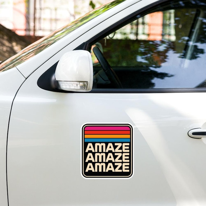 May include: A square car magnet featuring a retro-inspired design with horizontal stripes in pink, red, orange, and blue at the top. Below the stripes, the word AMAZE is printed in a bold, sans-serif font in three stacked rows on a cream background.