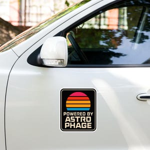 May include: A square car magnet featuring a graphic of a multi-colored sunset with horizontal stripes in pink, red, orange, and blue. Below the sunset, the text reads POWERED BY ASTRO PHAGE in a bold, sans-serif font against a black background.