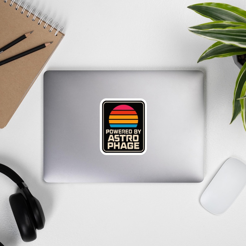 May include: A rectangular sticker with rounded corners featuring a retro-style sunset graphic in pink, orange, and blue horizontal stripes above the text POWERED BY ASTRO PHAGE in a bold, sans-serif font, all set against a solid black background.