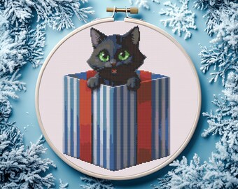 Pode incluir: Bordado em ponto cruz de um gato preto com olhos verdes espreitando de uma caixa de presente. A caixa é listrada em vermelho, branco e azul. O bordado está em um bastidor de madeira, em um fundo azul com detalhes de flocos de neve gelados.