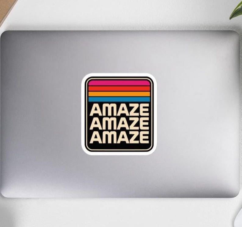 May include: A square sticker with rounded corners featuring a retro-style design. The top section has horizontal stripes in pink, red, orange, and blue. Below the stripes, the word AMAZE is printed in a bold, sans-serif font on three lines.