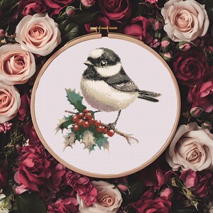 Könnte beinhalten: Kreuzstich-Stickerei eines kleinen Vogels auf einem Stechpalmenzweig mit roten Beeren. Der Vogel hat schwarze, weiße und beige Federn. Die Stickerei befindet sich in einem Holzreifen, umgeben von roten und rosa Rosen.