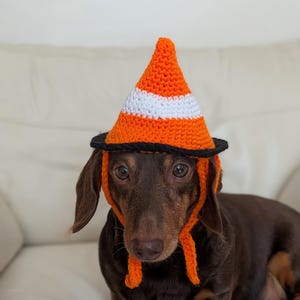 May include: A crochet hat shaped like a traffic cone, featuring a pointed orange top with a horizontal white stripe and a black brim. The hat includes orange ear flaps with dangling ties, designed to fit a small dog with brown fur and floppy ears.