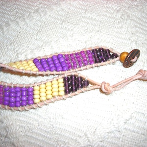 May include: A multi-colored beaded bracelet with purple, yellow, and brown beads. The bracelet is made of leather and has a brown wooden toggle clasp.
