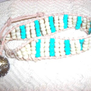 May include: A white and turquoise beaded bracelet with a silver charm. The bracelet is made of small, round beads that are strung together on a leather cord.