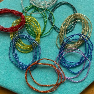 May include: A collection of colorful beaded waist beads in various shades of blue, green, red, orange, purple, and gold. The beads are arranged in a circular pattern on a turquoise surface.