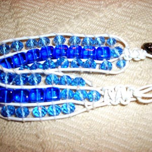 May include: A white cord bracelet with blue and light blue glass beads. The bracelet is woven with a knot pattern.