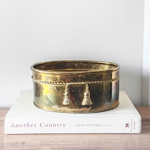 May include: A vintage brass oval planter with a rope detail and two tassels. The planter sits on a white book titled "Another Country" by Mary Pipher, Ph.D. The brass has a warm, aged patina.
