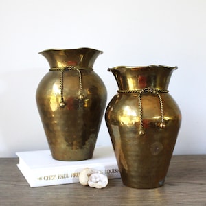 May include: Two vintage brass vases with a hammered texture and ruffled rims. Each vase has a decorative rope detail around the neck. The vases are displayed on a wooden surface with a book and small stones.